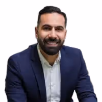 Smiling man with dark hair and beard wearing a navy blazer and white shirt against a white background.
