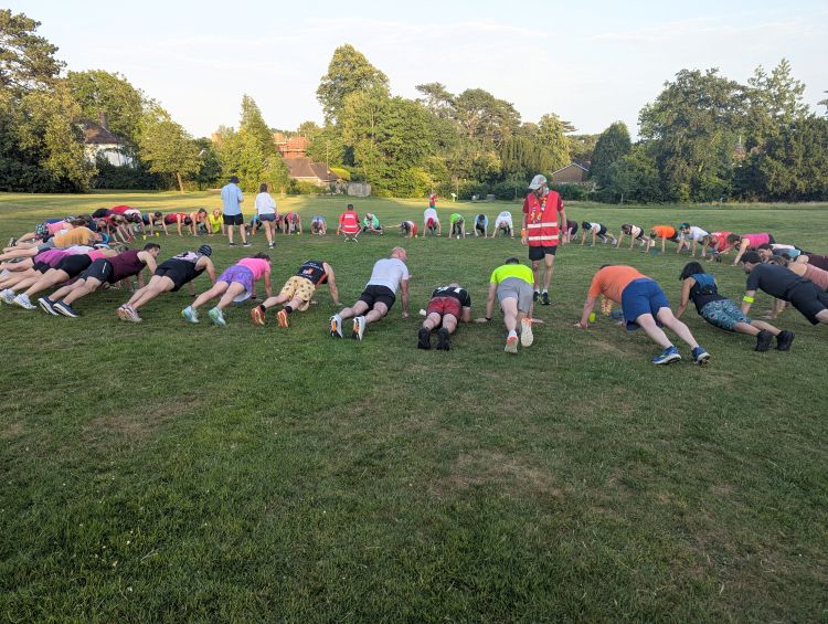 A large group holding a plank in a circle on a playing field, with a leader in a hi-vis vest