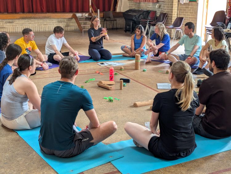 A workshop in a hall, with people sitting on mats in a circle around foam rollers and massage balls