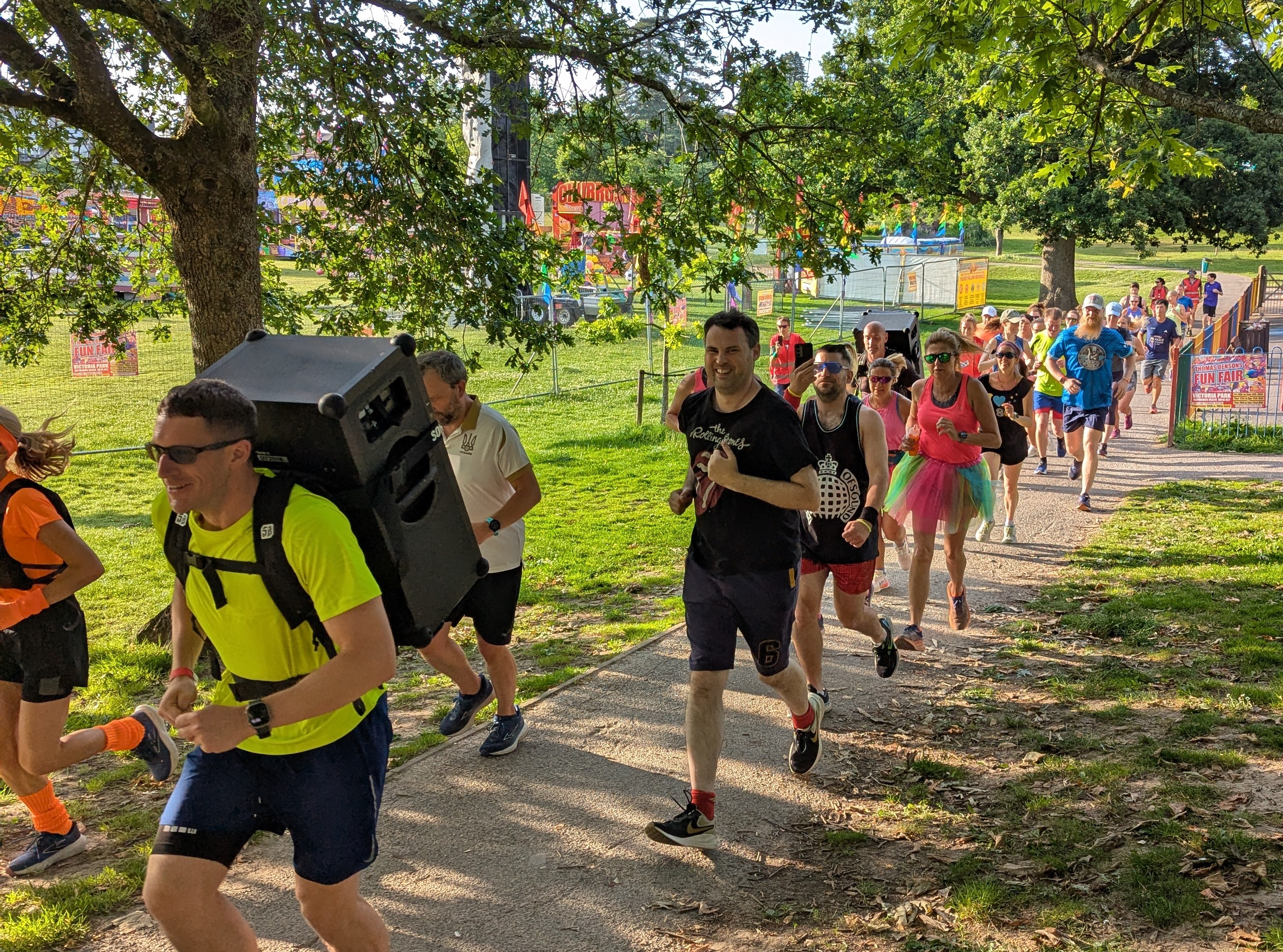 Runners setting off on the Rave Run through a sunny park, one carrying a large speaker on his back