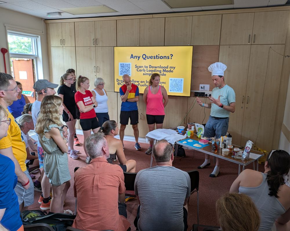 A cooking demonstration on carb loading, with the presenter in a chef’s hat and an audience gathered round