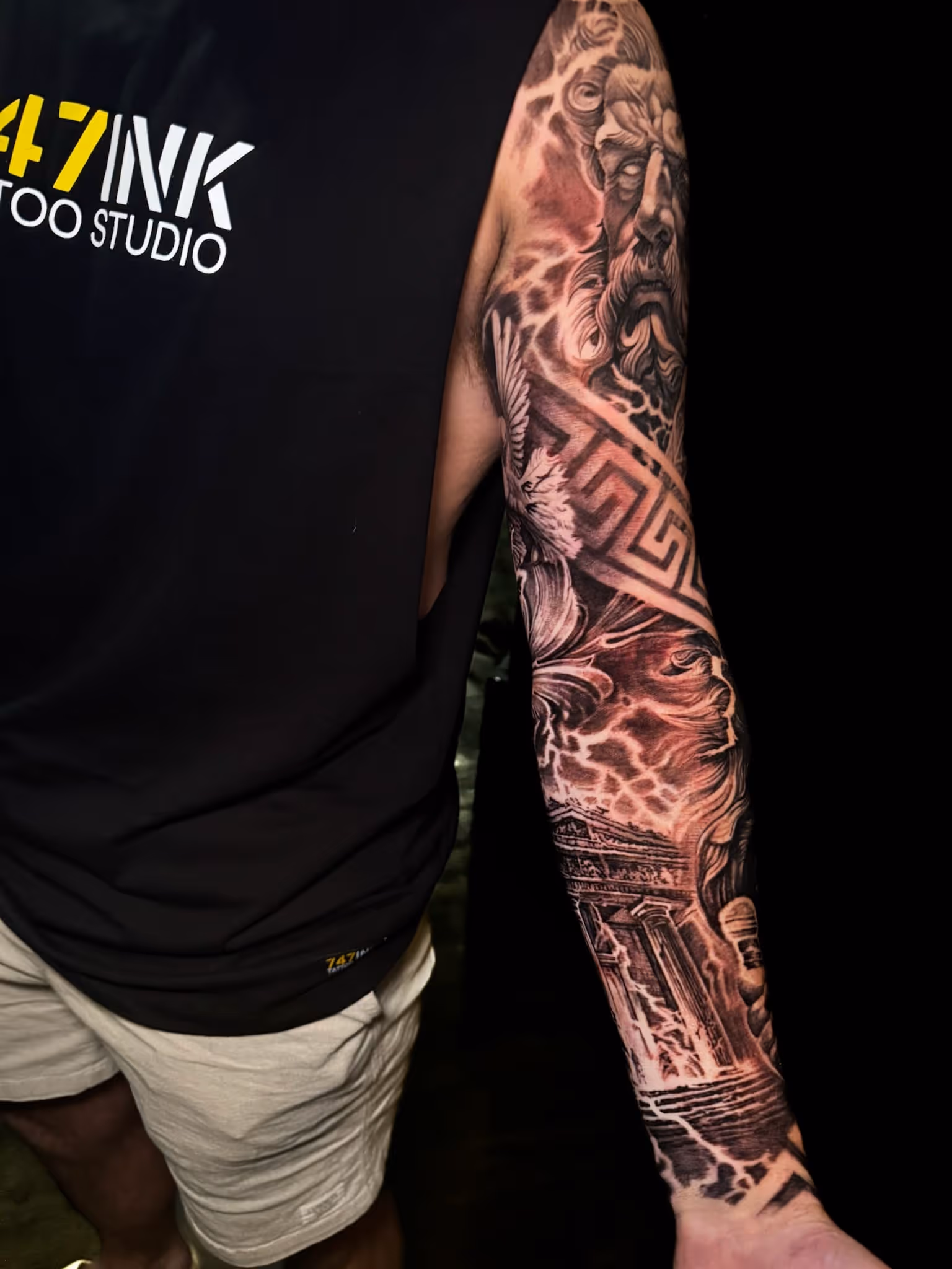 Detailed black and gray full-sleeve tattoo depicting an ancient Greek statue face, geometric patterns, an eagle, and a classical temple with lightning effects.