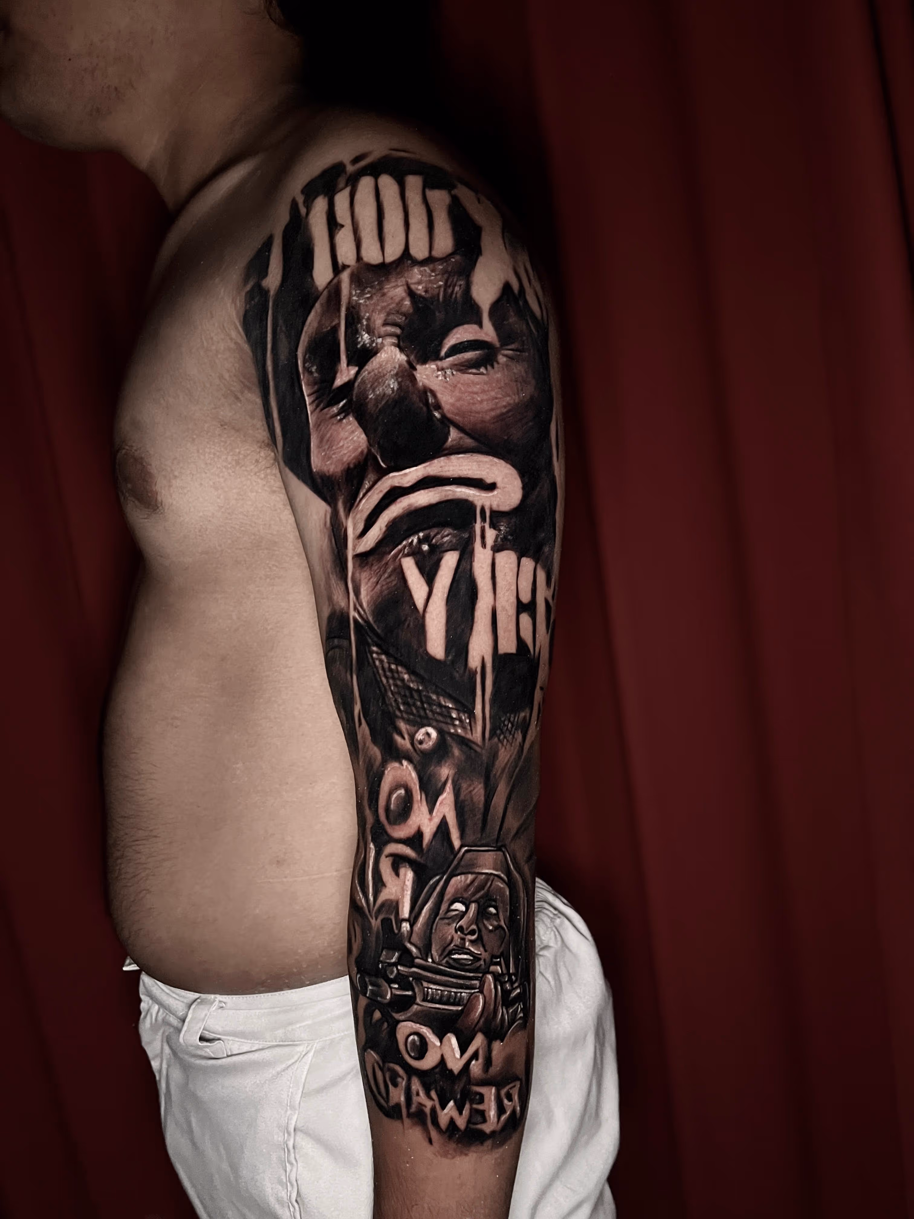 Black and grey tattoo sleeve on left arm depicting a sad face, a person wearing a helmet with a rifle, and stylized text elements.