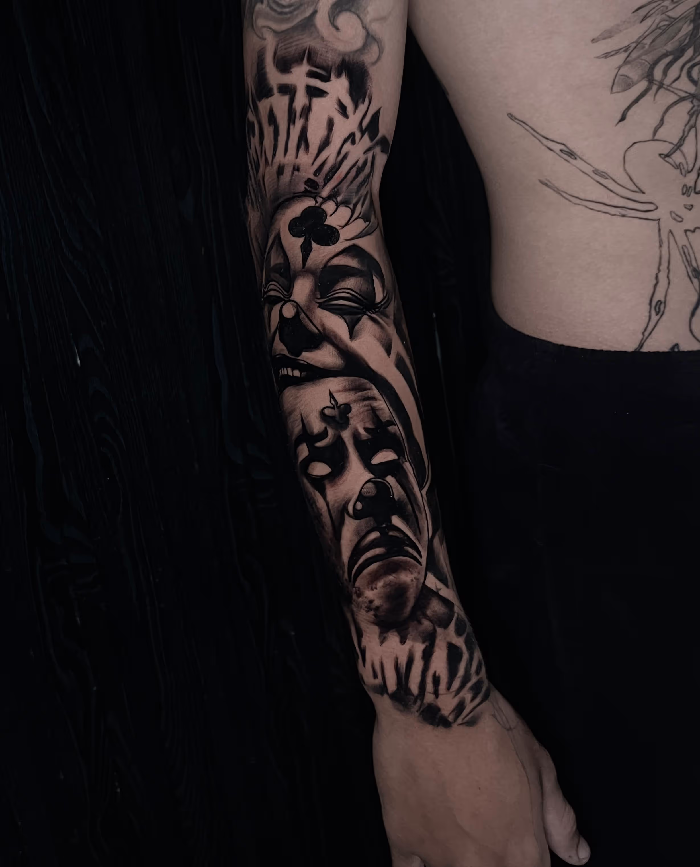 Black and grey tattoo of two theatrical clown faces, one laughing and one crying, on a person's forearm.
