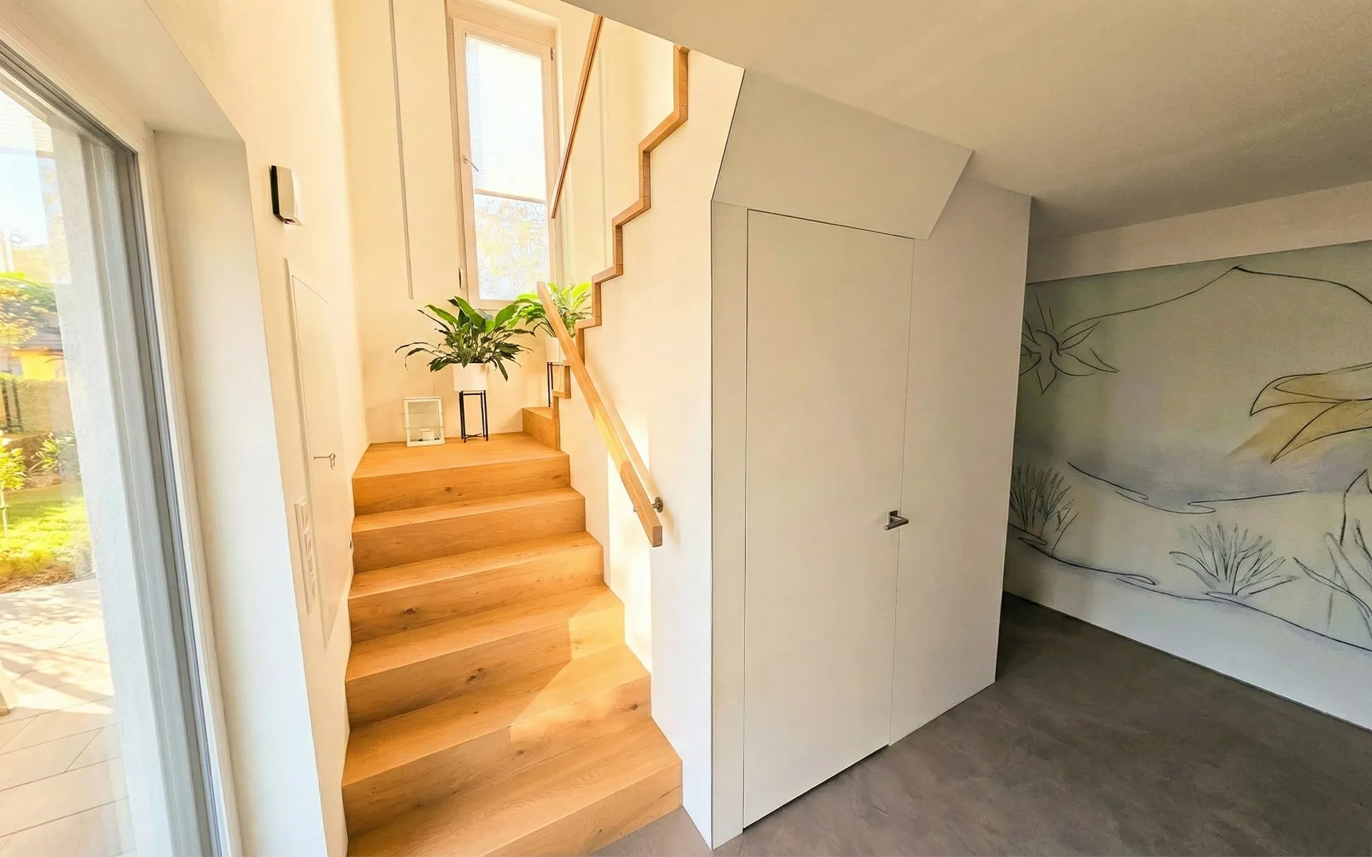 Helle Holztreppe mit Pflanzen und Fenstern neben einer weißen Wand mit Tür und Wandkunst.