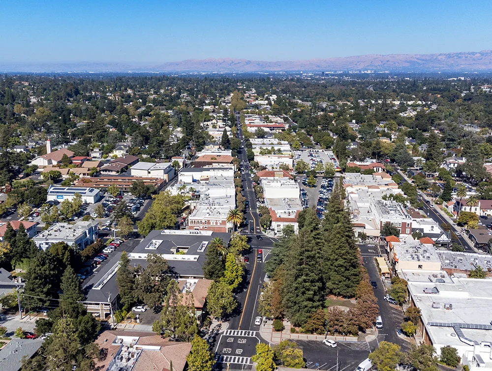 Aerial view of downtown Los Gatos, California with a mix of commercial buildings and residential homes. The green suburban landscape extends toward distant mountains