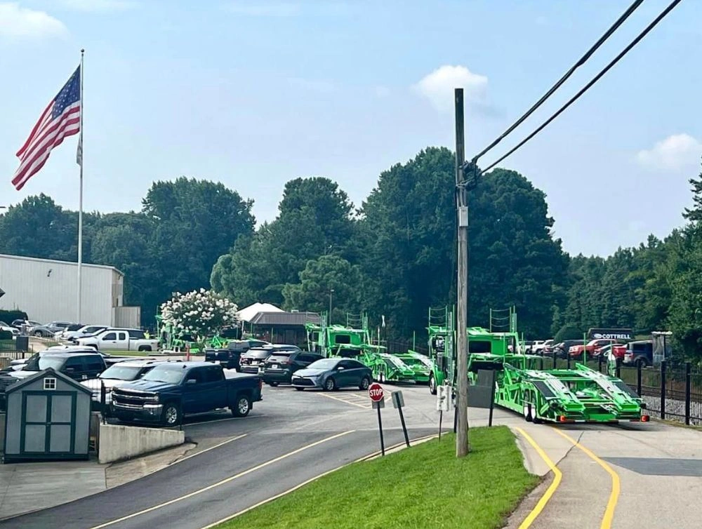 Entrance road to a truck and trailer facility with green car carriers, parked vehicles, utility poles, and an American flag