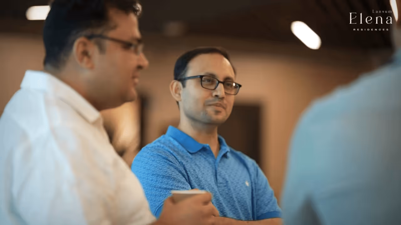 Three men engaged in a casual conversation indoors, one wearing a blue polo shirt and glasses holding a cup.
