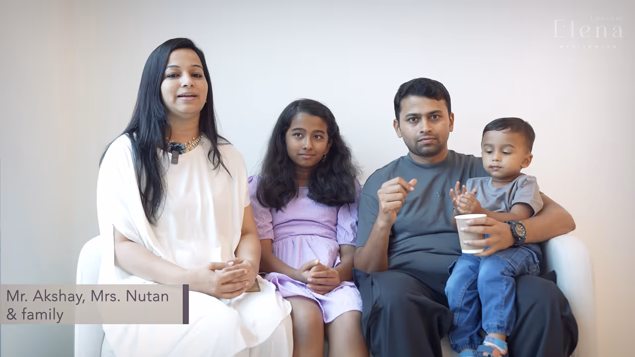 A family of four sitting on a white couch against a plain wall; a woman in a white dress, a girl in a purple dress, a man in a gray shirt holding a young boy with a cup.