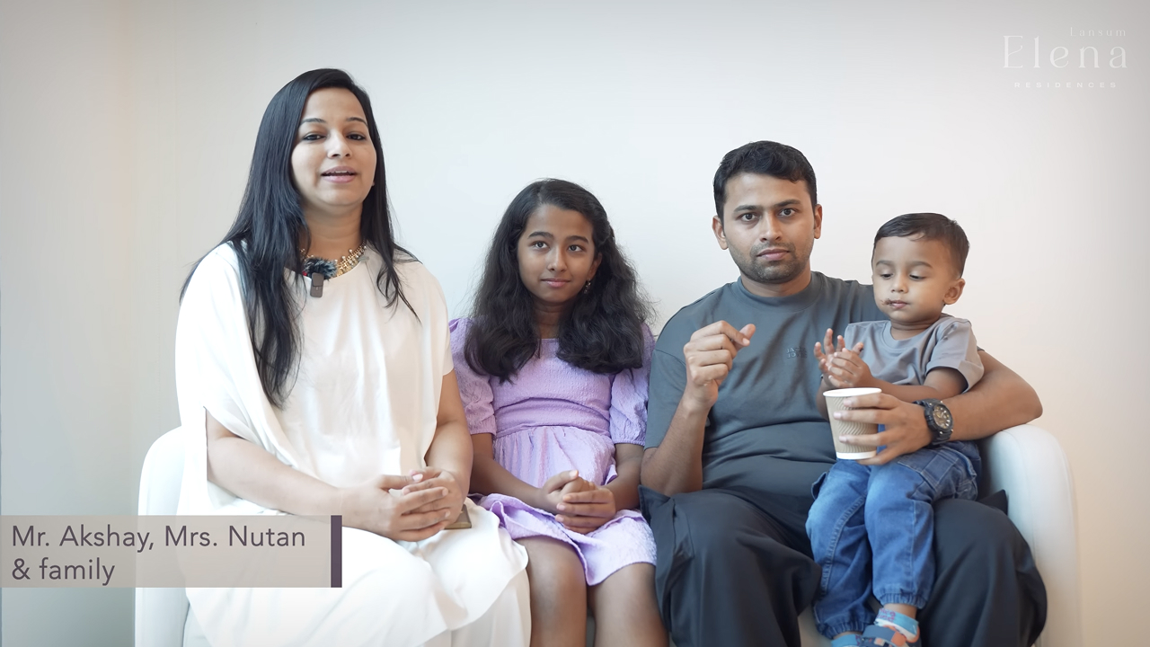 A family of four sitting on a white couch against a plain wall; a woman in a white dress, a girl in a purple dress, a man in a gray shirt holding a young boy with a cup.