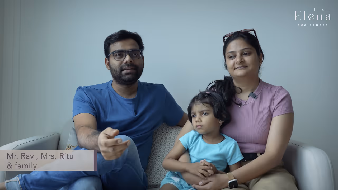A family of three sitting on a sofa, with a man in a blue shirt, a woman in a pink shirt holding a young girl in a blue dress.