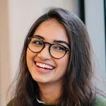 Smiling woman with long dark hair and glasses wearing a dark top against a light background.