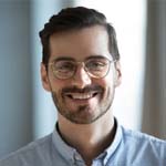 Smiling man with glasses, beard, and short dark hair wearing a light blue shirt in front of a blurred background.