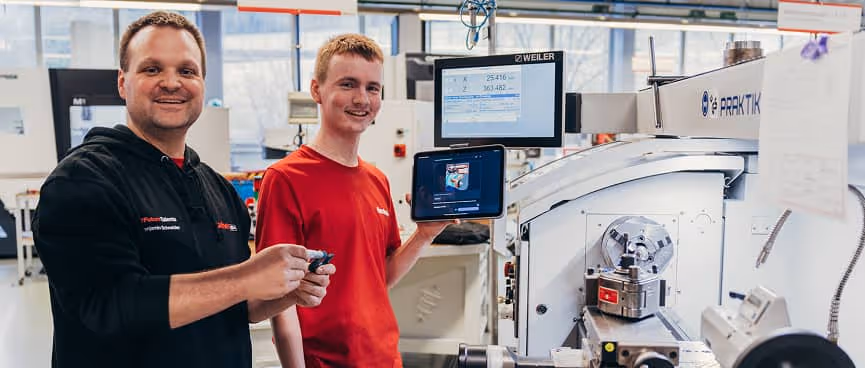 Ausbilder und Azubi lächeln in einer Werkstatt. Azubi hält Tablet mit Lerninhalten vor einer Maschine. Zeigt praktisches Lernen mit digitaler Unterstützung.