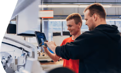 Ausbilder und Azubi schauen gemeinsam auf ein Tablet in einer Werkstatt. Zeigt Nutzung digitaler Lerninhalte für die Praxis.