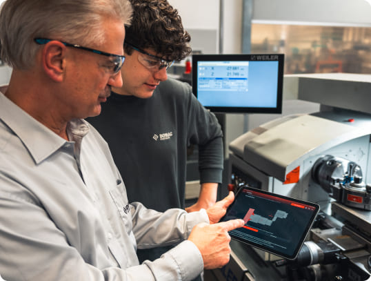 Ein Ausbilder und ein Auszubildender schauen auf ein Tablet mit einer technischen Zeichnung/Messung, während sie an einer CNC-Maschine stehen. Demonstriert digitale Fachkräfteausbildung im technischen Bereich