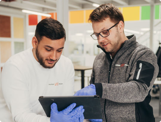 Zwei Männer (Maler oder Lackierer) mit Schutzhandschuhen schauen auf ein Tablet. Zeigt digitale Schulung und Anleitung im Handwerk.