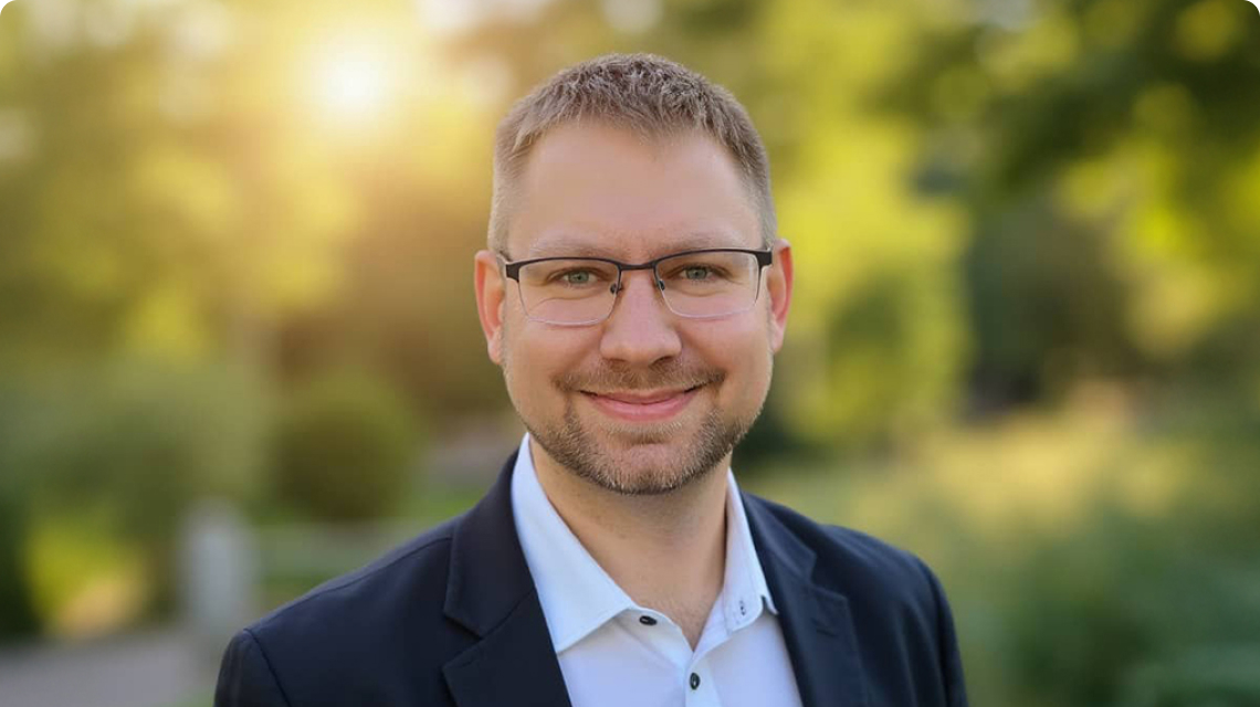 Mitarbeiter von KLA Tencor steht im Freien vor unscharfem Hintergrund, trägt Brille und Anzug und lächelt in die Kamera.