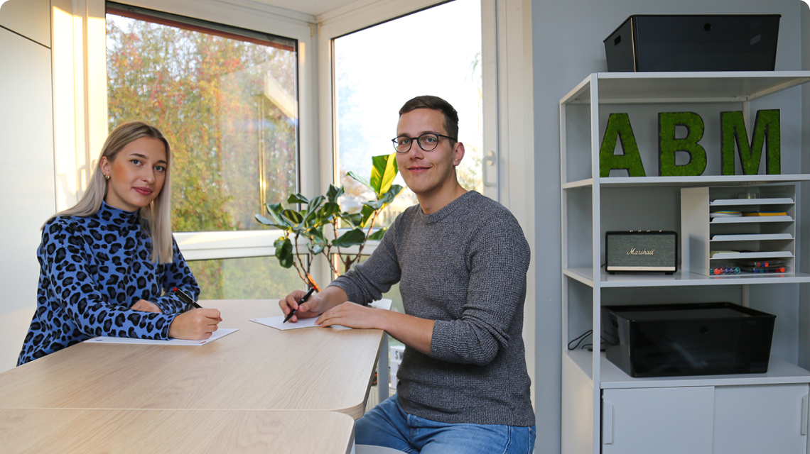 Mitarbeiter von ABM Technik Service sitzt am Schreibtisch vor einem Fenster, hält einen Stift in der Hand und lächelt in die Kamera.