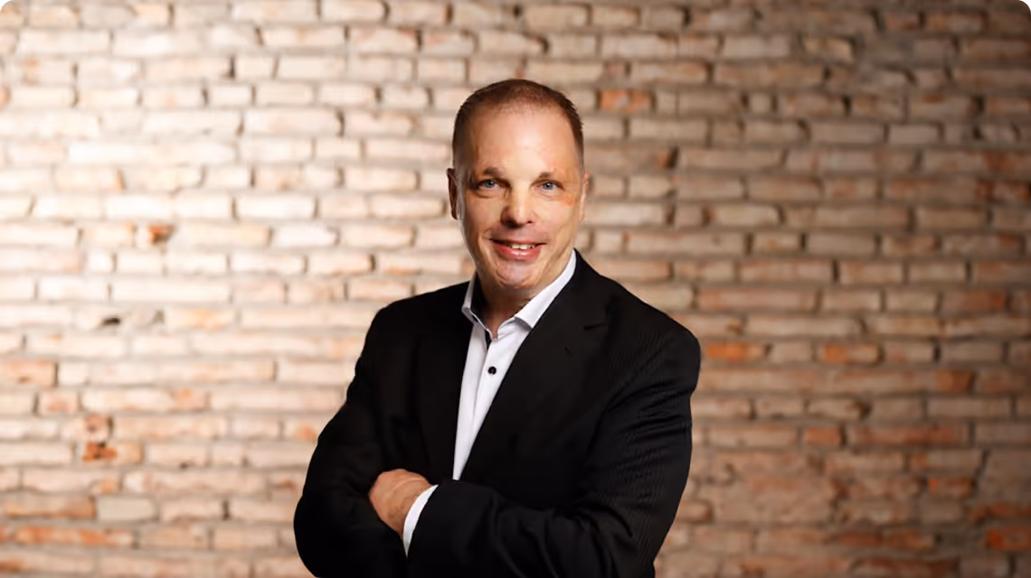 Mitarbeiter von Scheuch steht vor einer Backsteinwand, verschränkt die Arme und lächelt in die Kamera.