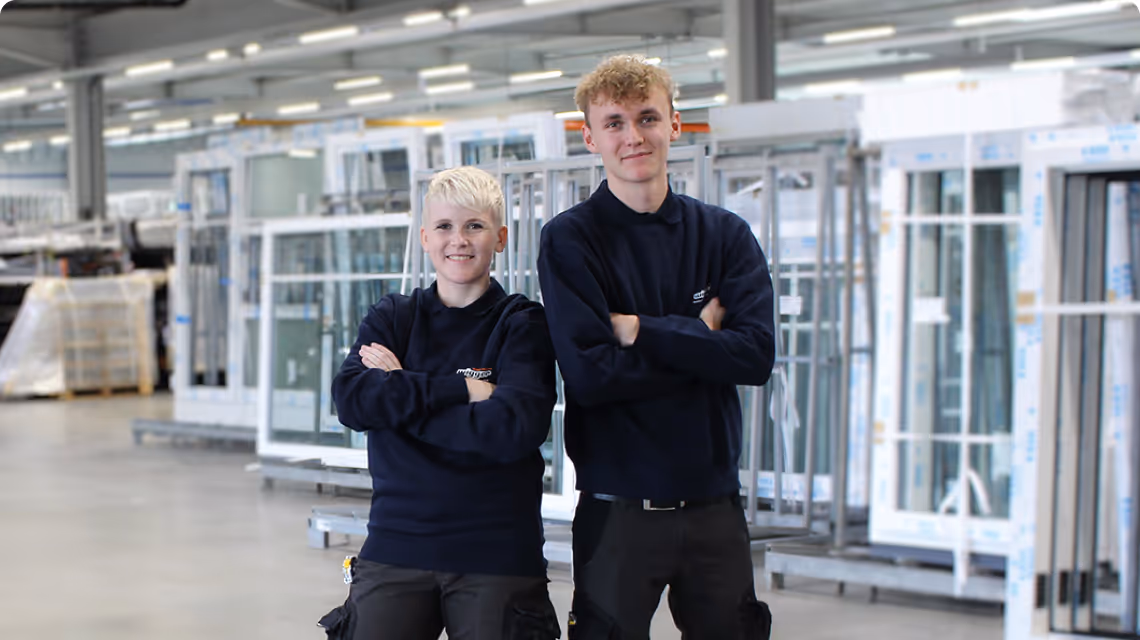 Zwei Auszubildende bei Wigger stehen mit verschränkten Armen in einer Produktionshalle und lächeln in die Kamera.