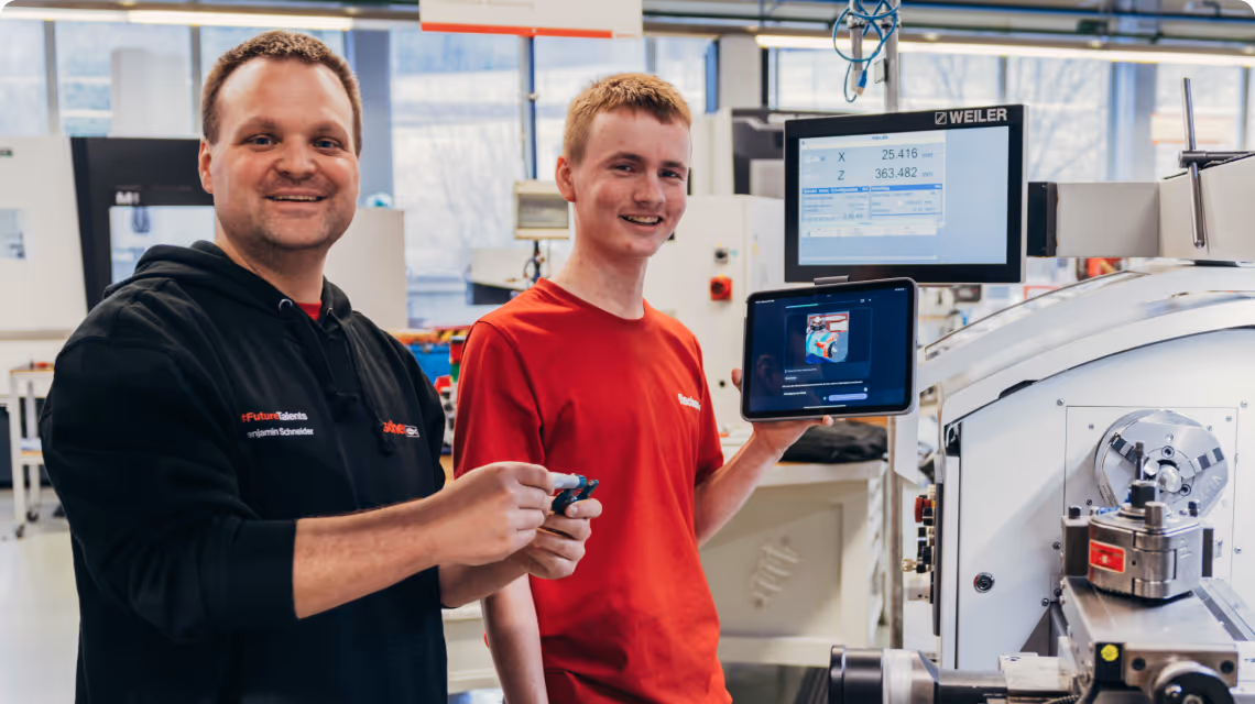 Zwei Auszubildende bei Fischer arbeiten an einem Werkstück und nutzen ein Tablet mit simpleclub zur Unterstützung.