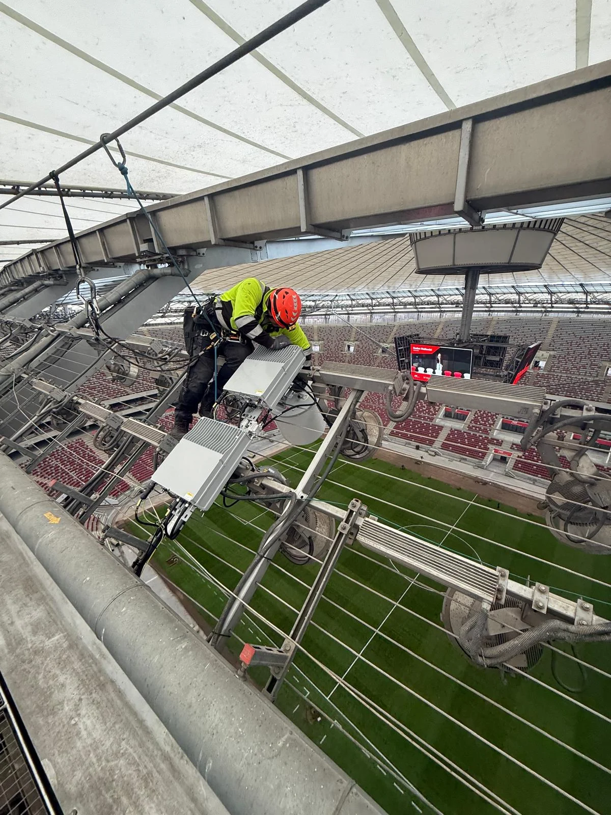 Pracownik w kasku i odblaskowej koszulce montuje sprzęt telekomunikacyjny na konstrukcji stadionu PGE Narodowy.
