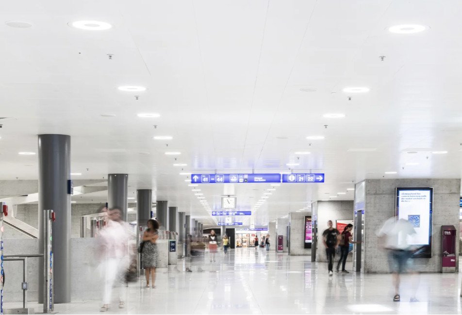 Wnętrze nowoczesnego dworca kolejowego z dużą otwartą przestrzenią, biało-szarymi kolorami i rozmazanymi postaciami ludzi