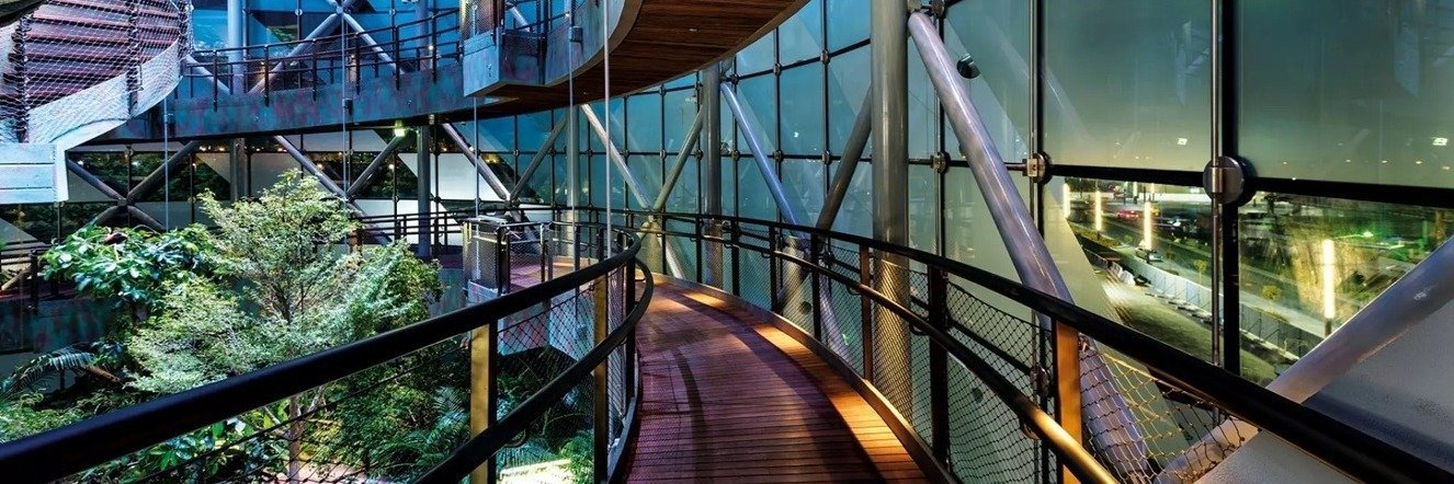 Architektura wnętrza ze szklanym dachem i drewnianymi kładkami połączonymi metalowymi siatkami, otoczonymi zielenią