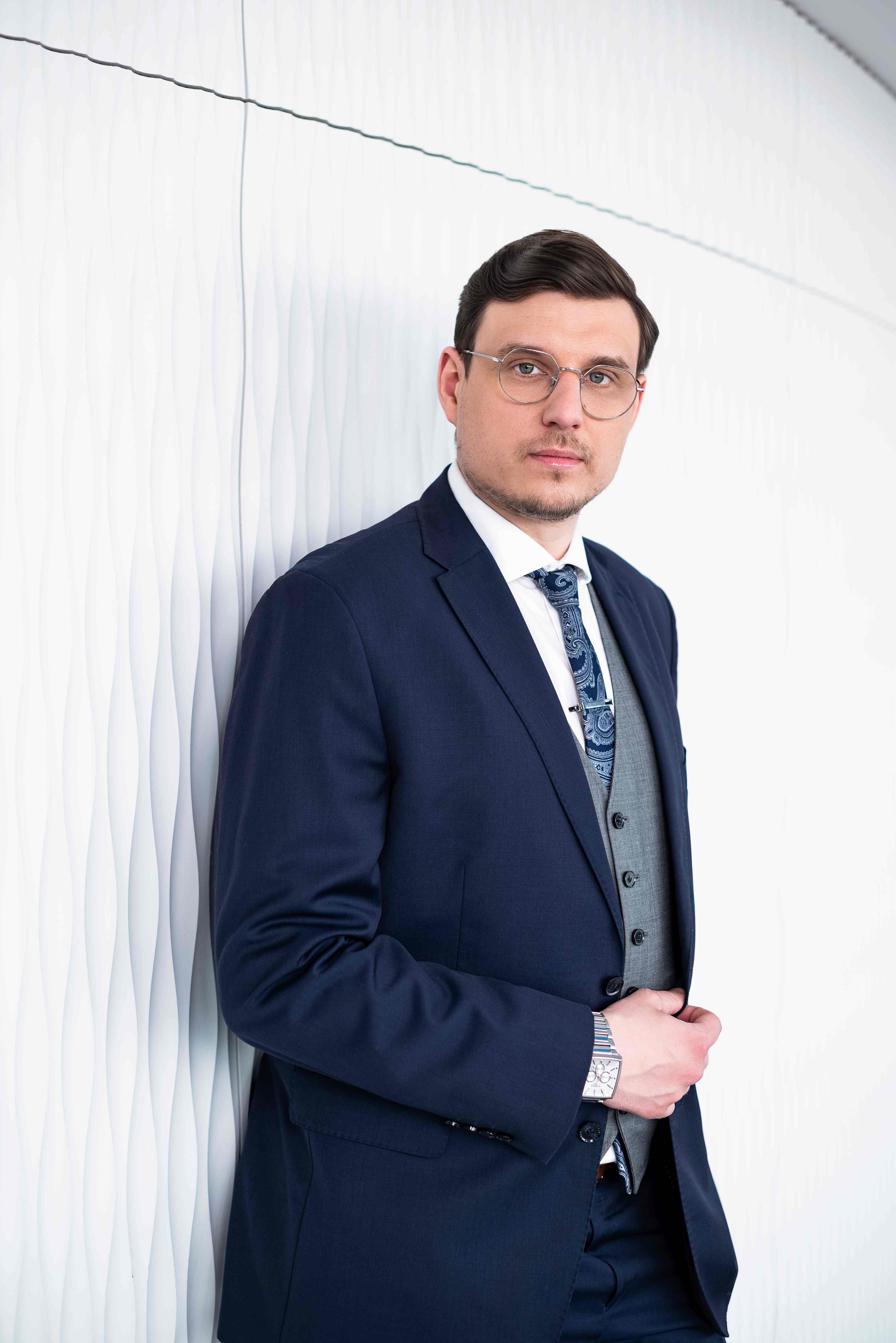 Mężczyzna w okularach i granatowym garniturze na tle białej dekoracyjnej ściany