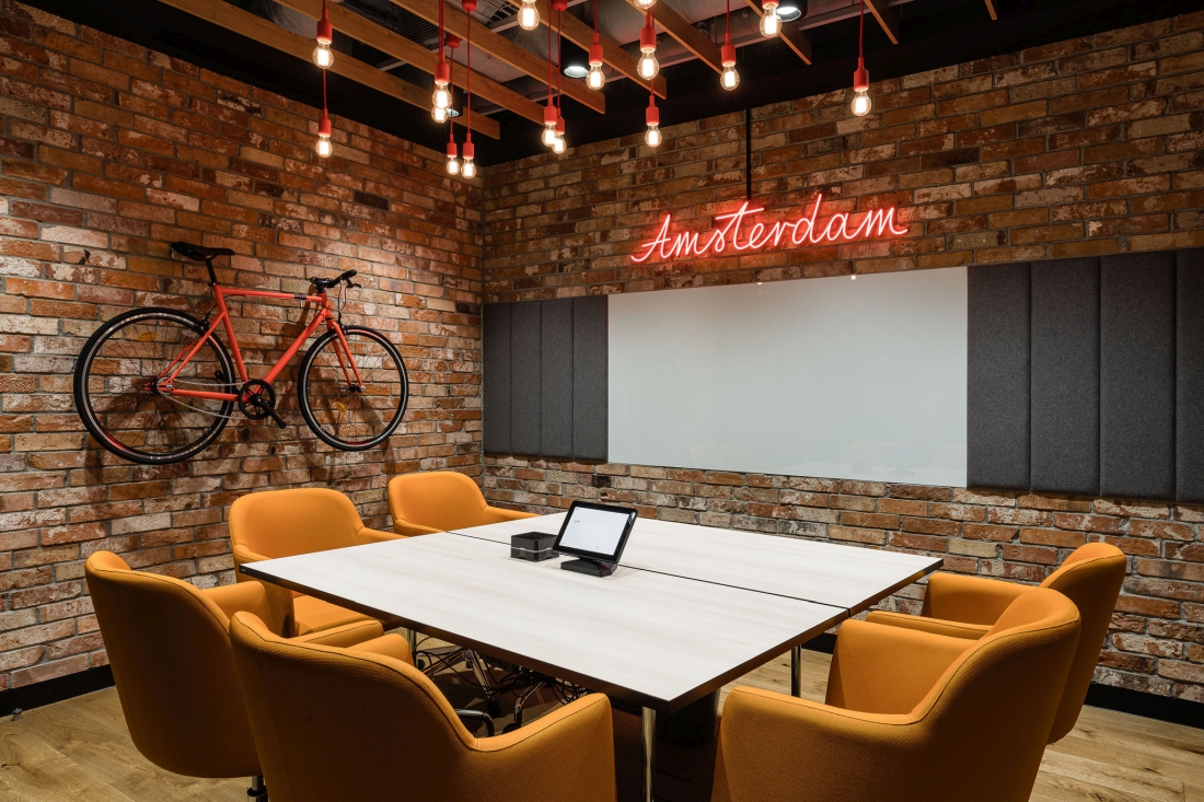 Nowoczesna sala konferencyjna o industrialnym charakterze z czerwonymi ceglanymi ścianami, pomarańczowym rowerem, neonowym napisem „Amsterdam” i białym stołem z musztardowymi fotelami.
