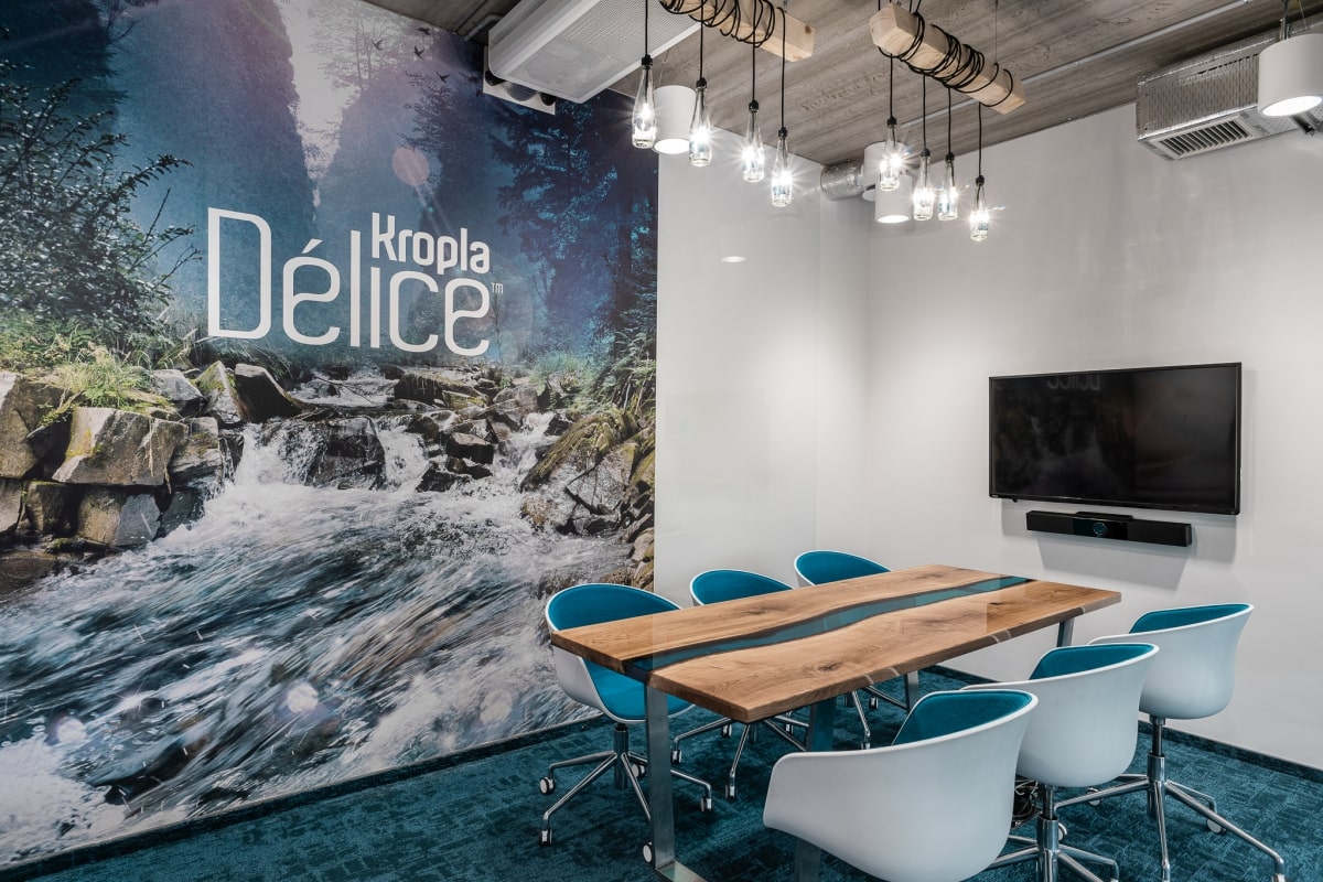 Nowoczesna sala konferencyjna z drewnianym stołem z błękitnym przeszkleniem typu „river table” i biało-niebieskimi fotelami. Na ścianie fototapeta przedstawiająca górski potok i logotyp „Kropla Délice”
