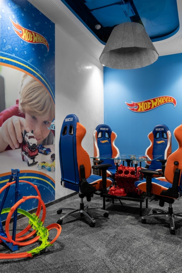 Pomieszczenie biurowe lub salka konferencyjna urządzona w motoryzacyjnym stylu z logo Hot Wheels na niebieskich ścianach i fototapecie, ze szklanym stolikiem opartym na czerwonym korpusie silnika.