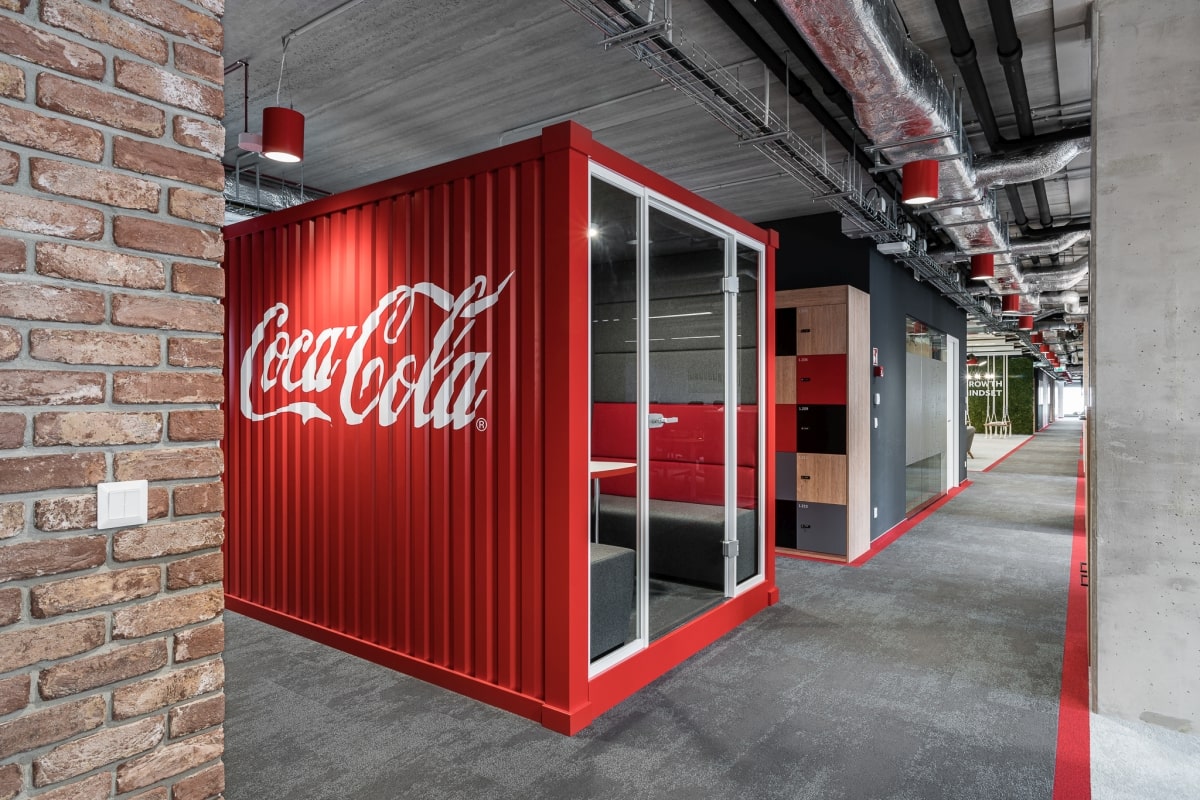 Nowoczesne wnętrze biurowe w stylu industrialnym z czerwoną budką telefoniczną lub salką spotkań imitującą kontener transportowy z dużym logo Coca-Cola.