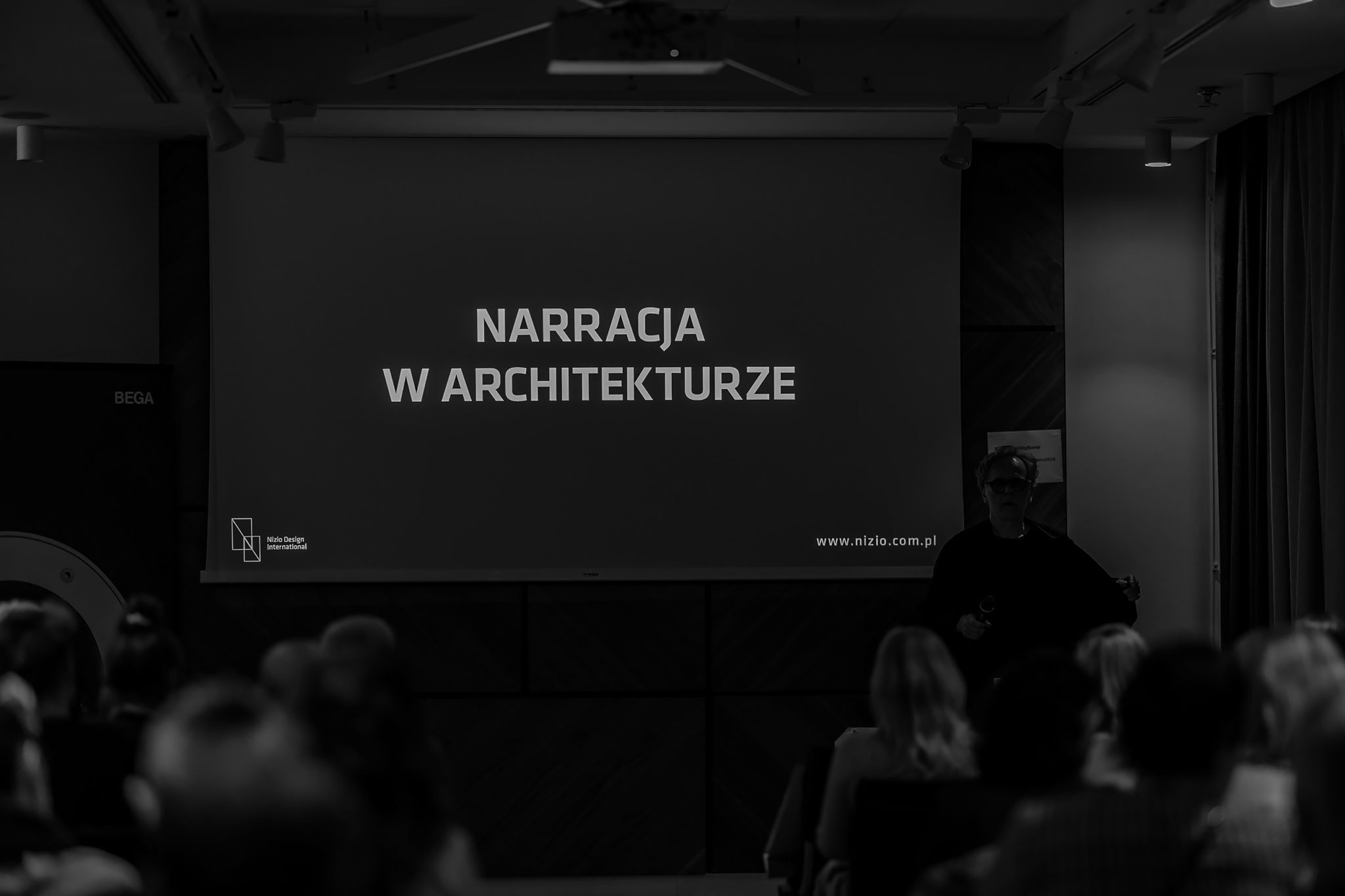 Czarno-biała fotografia sali wykładowej z prelegentem i prezentacją 'NARRACJA W ARCHITEKTURZE'