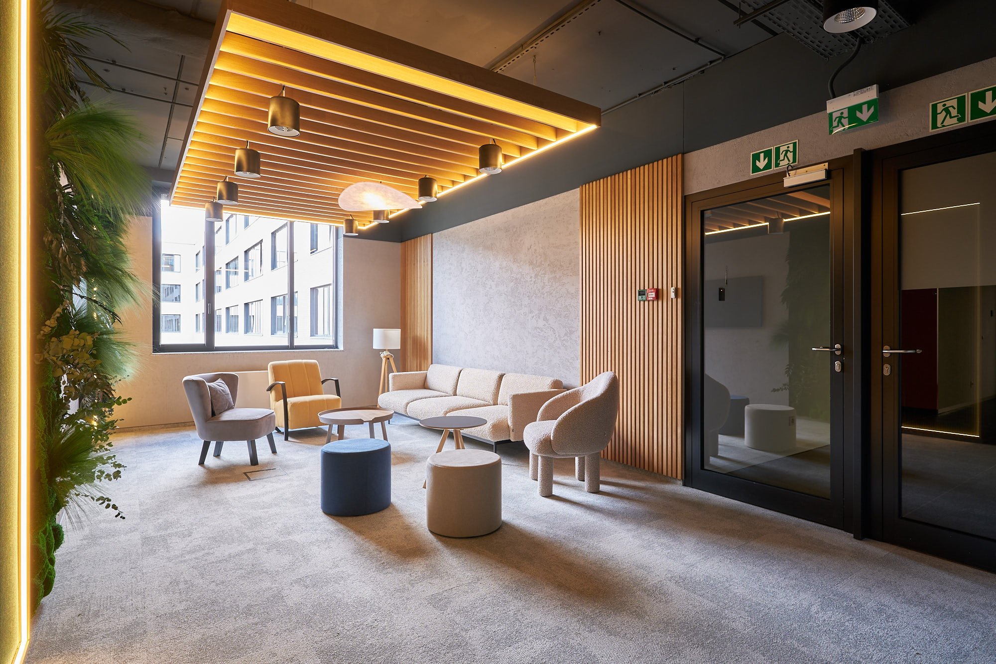 Nowoczesna strefa wypoczynkowa z tapicerowaną sofą, fotelami, pufami i panelami ściennymi z drewna oraz betonu architektonicznego.