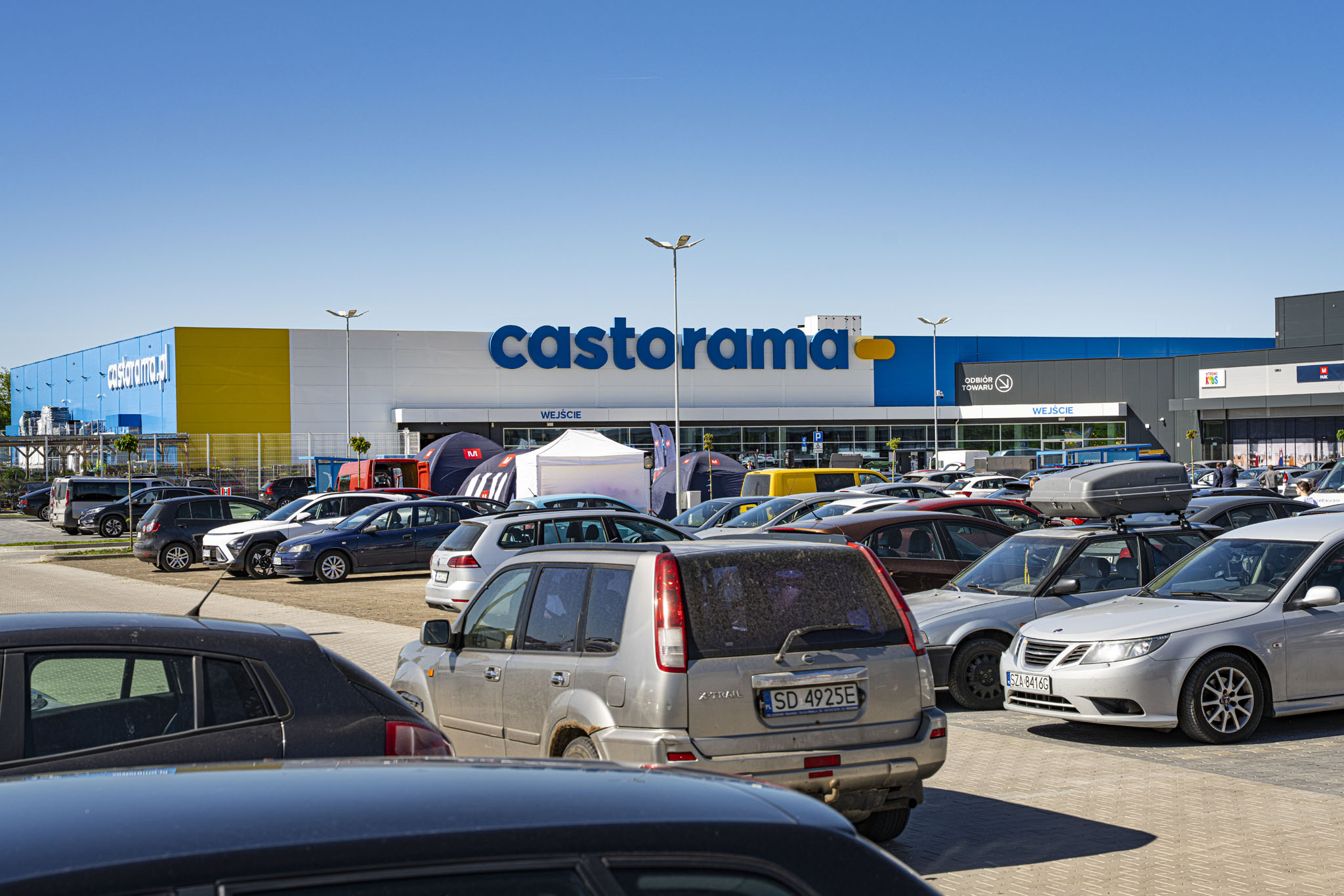 Wielkopowierzchniowy sklep budowlany Castorama o biało-niebiesko-żółtej elewacji z parkingiem pełnym samochodów.
