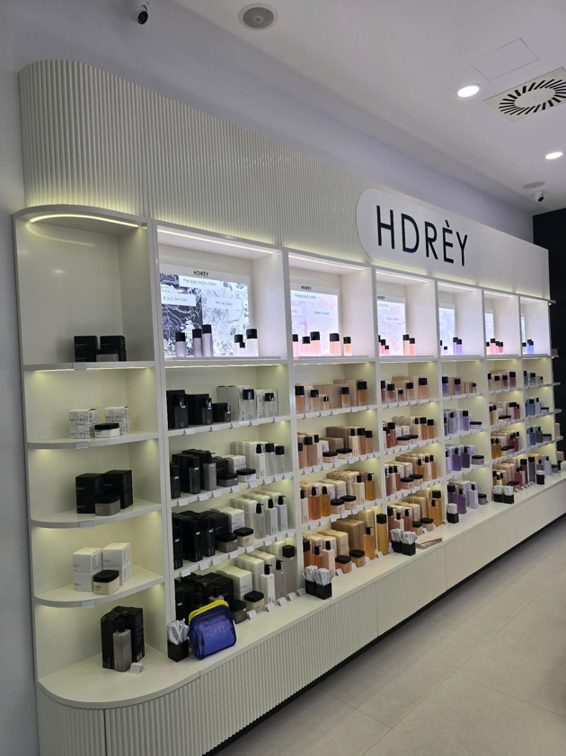 Nowoczesny regał sklepowy z kosmetykami marki HDREY w minimalistycznym wnętrzu, z białymi podświetlanymi półkami z flakonami perfum i produktami do pielęgnacji.