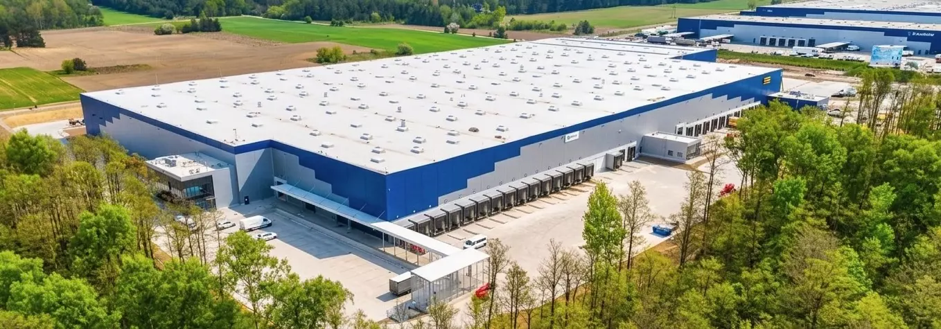 Wielkopowierzchniowa hala magazynowo-logistyczna o płaskim dachu z licznymi świetlikami, z elewacją w kolorach jasnoszarym i niebieskim, otoczona utwardzonymi placami manewrowymi.