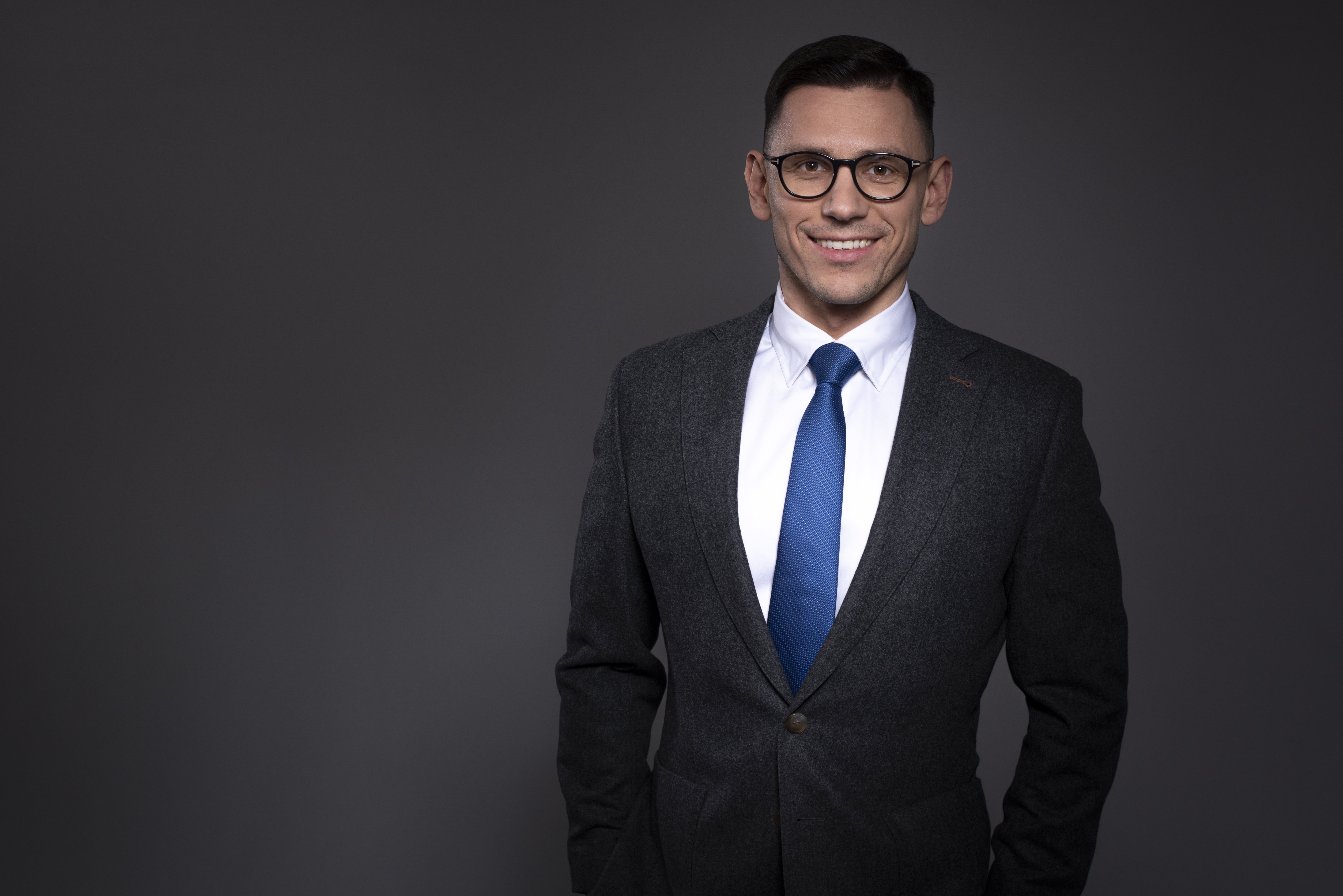 Mężczyzna w średnim wieku, noszący okulary, uśmiecha się patrząc w obiektyw, ubrany w szary garnitur i niebieski krawat.