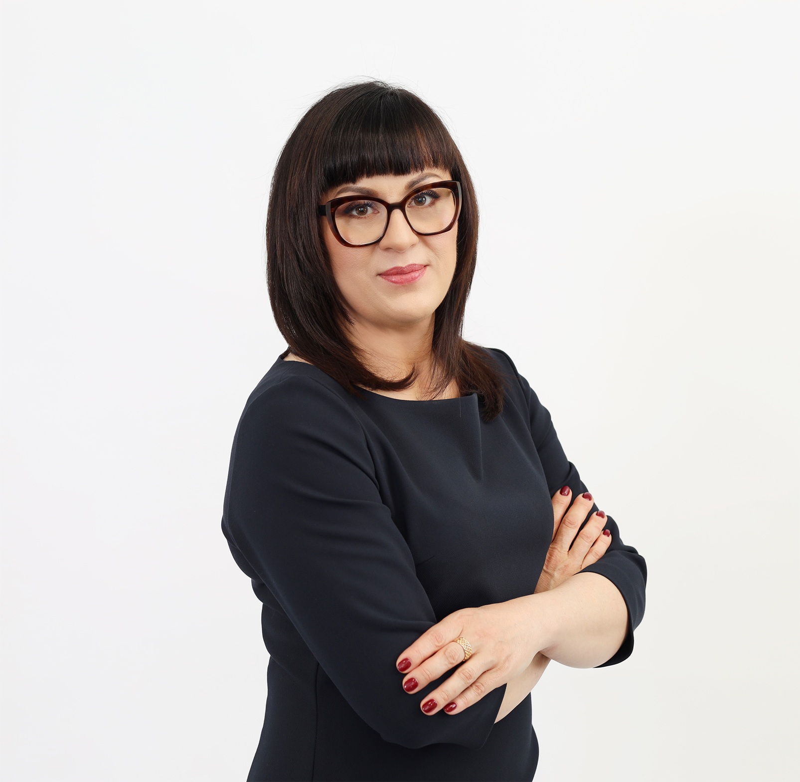 Portret kobiety o ciemnych włosach z prostą grzywką, stojącej na jednolitym, białym tle w pozie z założonymi rękami.