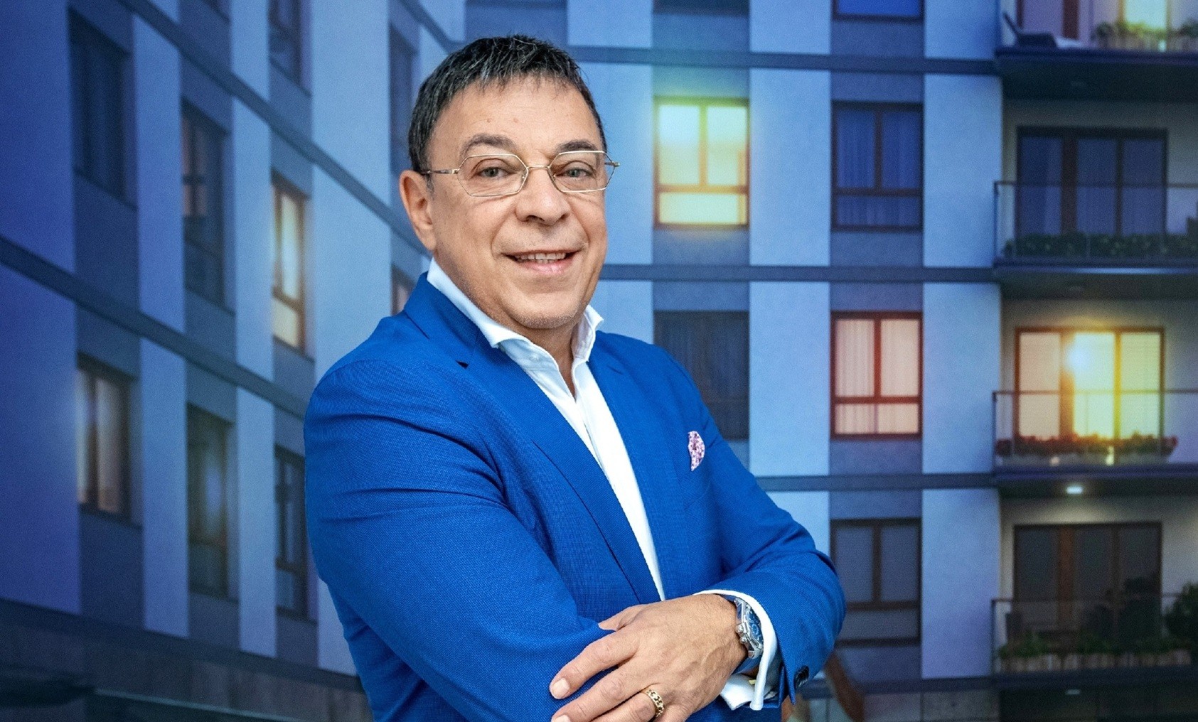 Portret uśmiechniętego mężczyzny w średnim wieku, ubranego w jaskrawoniebieską marynarkę, białą koszulę i okulary korekcyjne, pozującego z założonymi rękami.