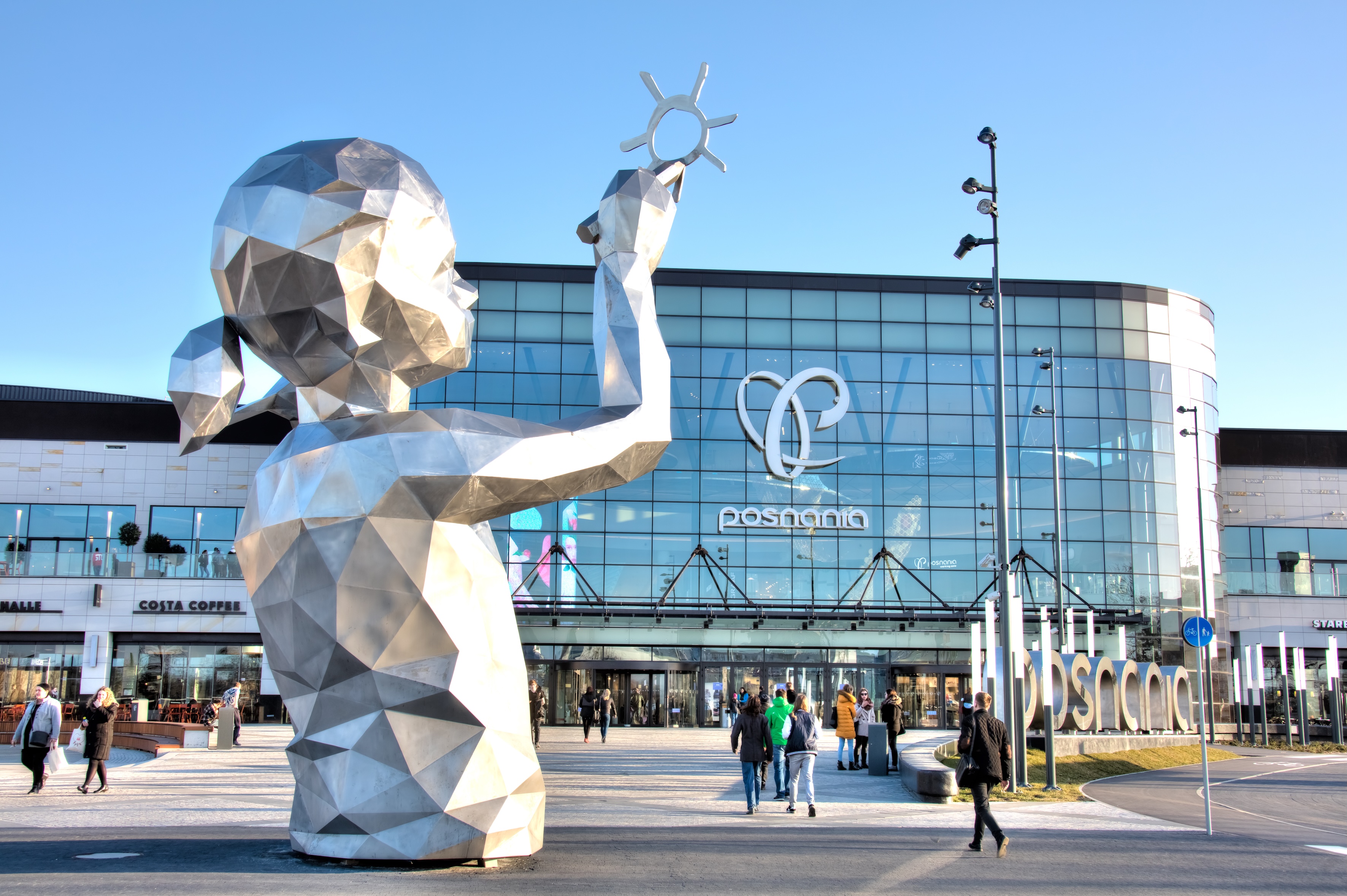 Przed nowoczesnym budynkiem centrum handlowego Posnania, stoi srebrna rzeźba dziecka z symbolem słońca.