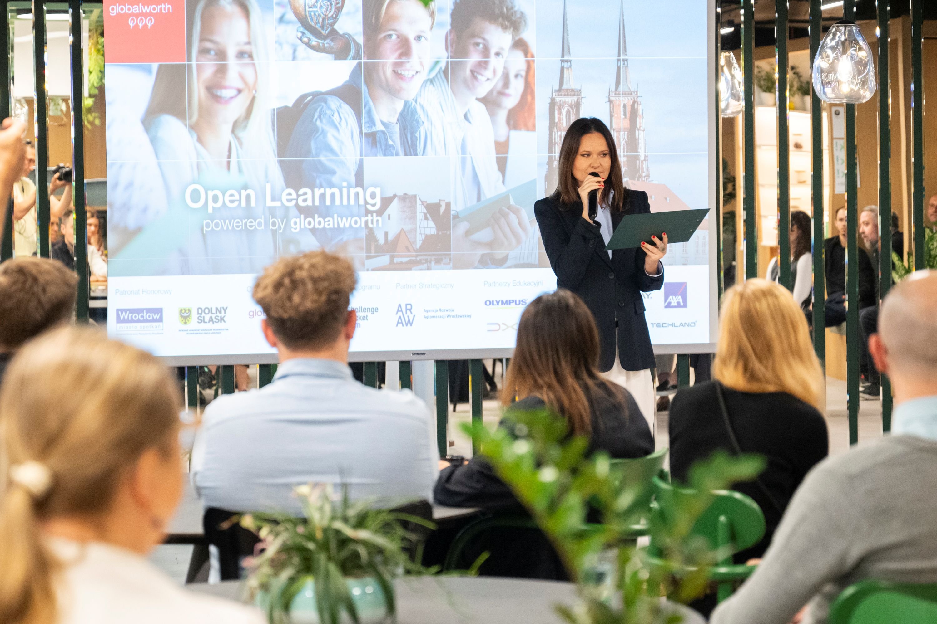 Kobieta w ciemnej marynarce przemawia do mikrofonu przed ekranem z napisem „Open Learning” i logotypami partnerów.