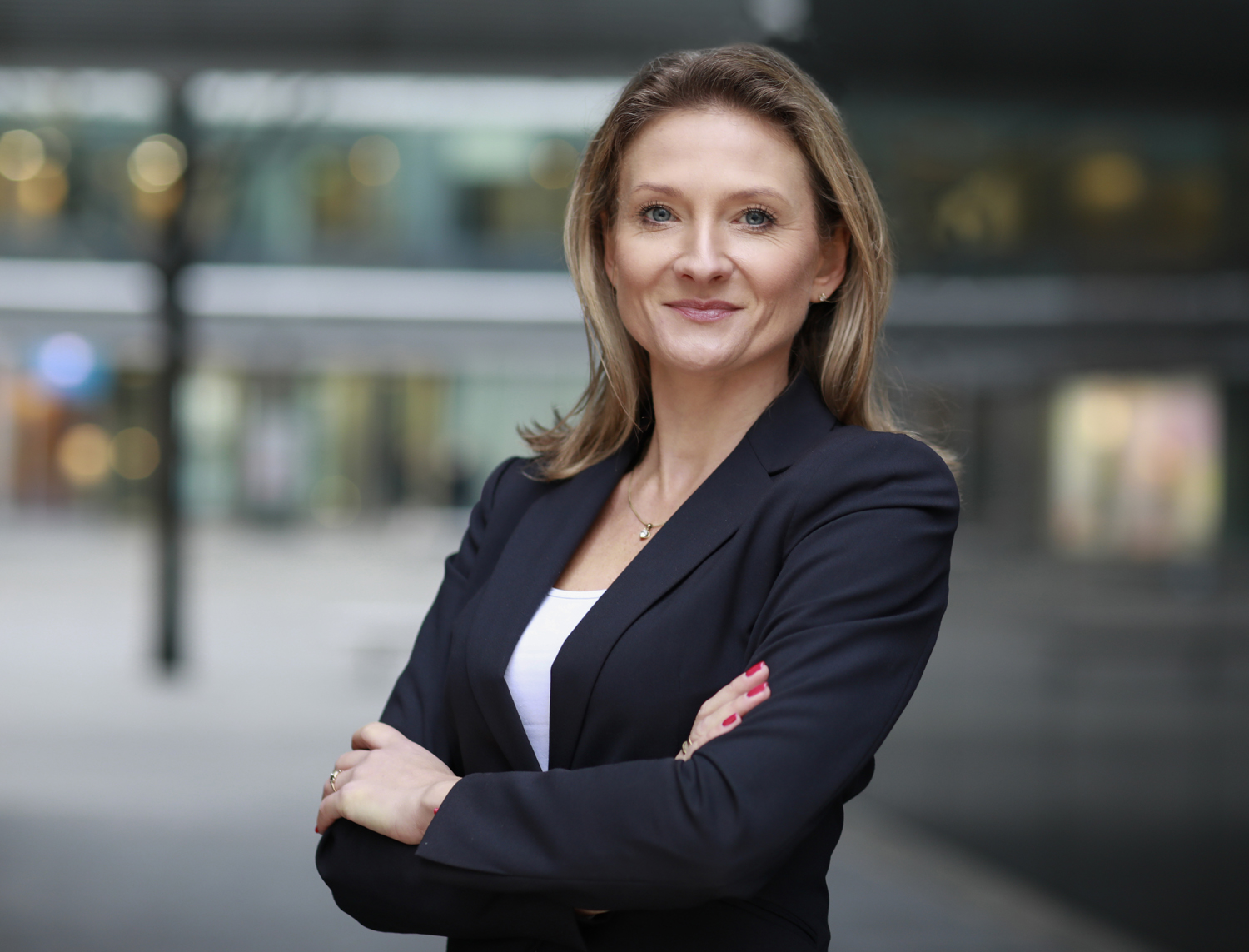 Portret uśmiechniętej kobiety o blond włosach z założonymi rękami, ubranej w ciemną marynarkę na tle rozmytego, miejskiego otoczenia.