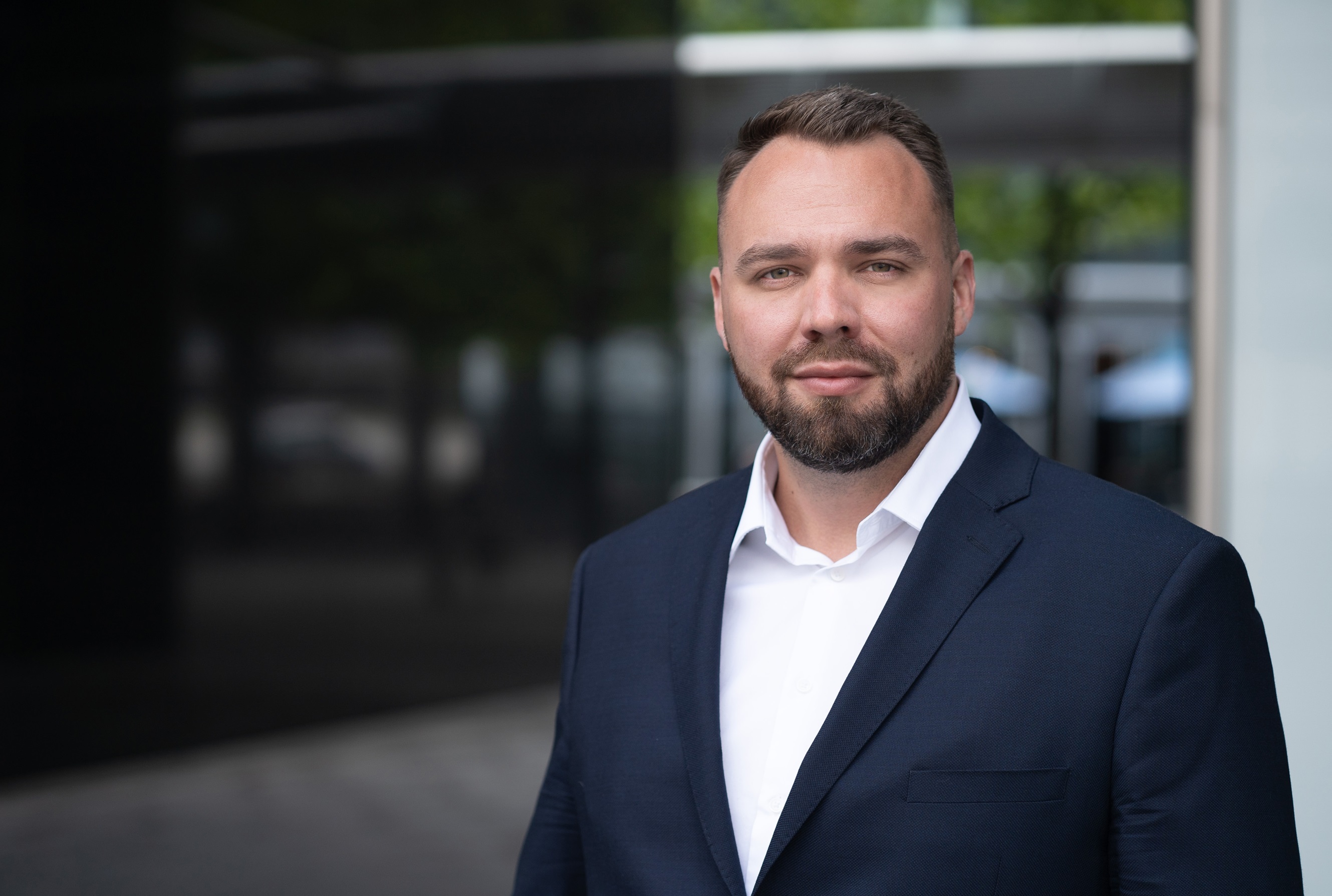 Portret mężczyzny z krótkim zarostem, w białej koszuli i ciemnogranatowej marynarce, patrzącego w obiektyw z lekkim uśmiechem.