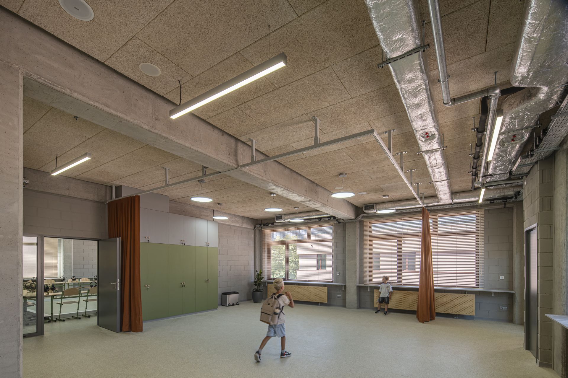 Nowoczesna, przestronna sala szkolna o industrialnym wykończeniu z betonowymi belkami i rurami wentylacyjnymi.