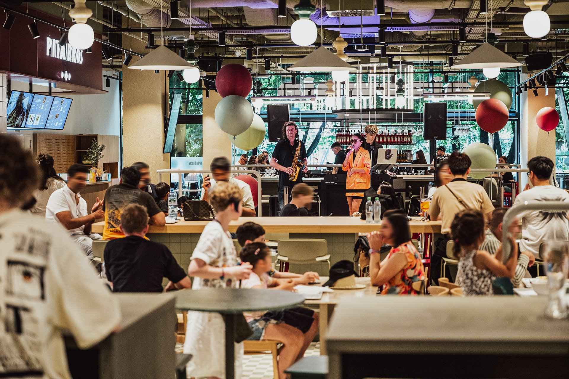 Tętniący życiem lokal gastronomiczny o nowoczesnym, industrialnym wystroju z licznymi gośćmi, sceną z saksofonistą i wokalistką.