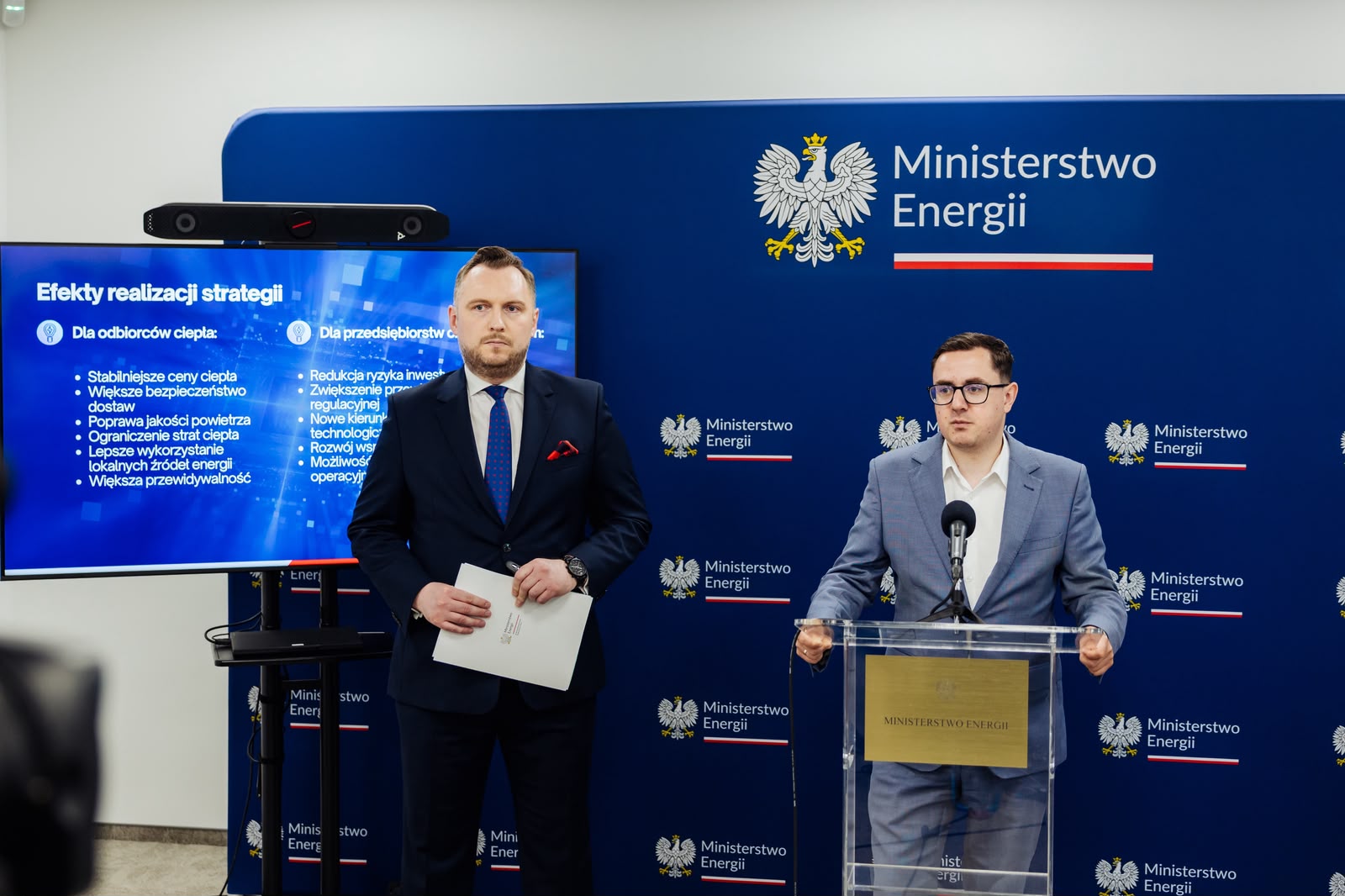 Dwaj mężczyźni w garniturach podczas konferencji prasowej w Ministerstwie Energii. Po prawej minister Motyka przy mównicy, po lewej minister Wojnarowski trzymający dokumenty.