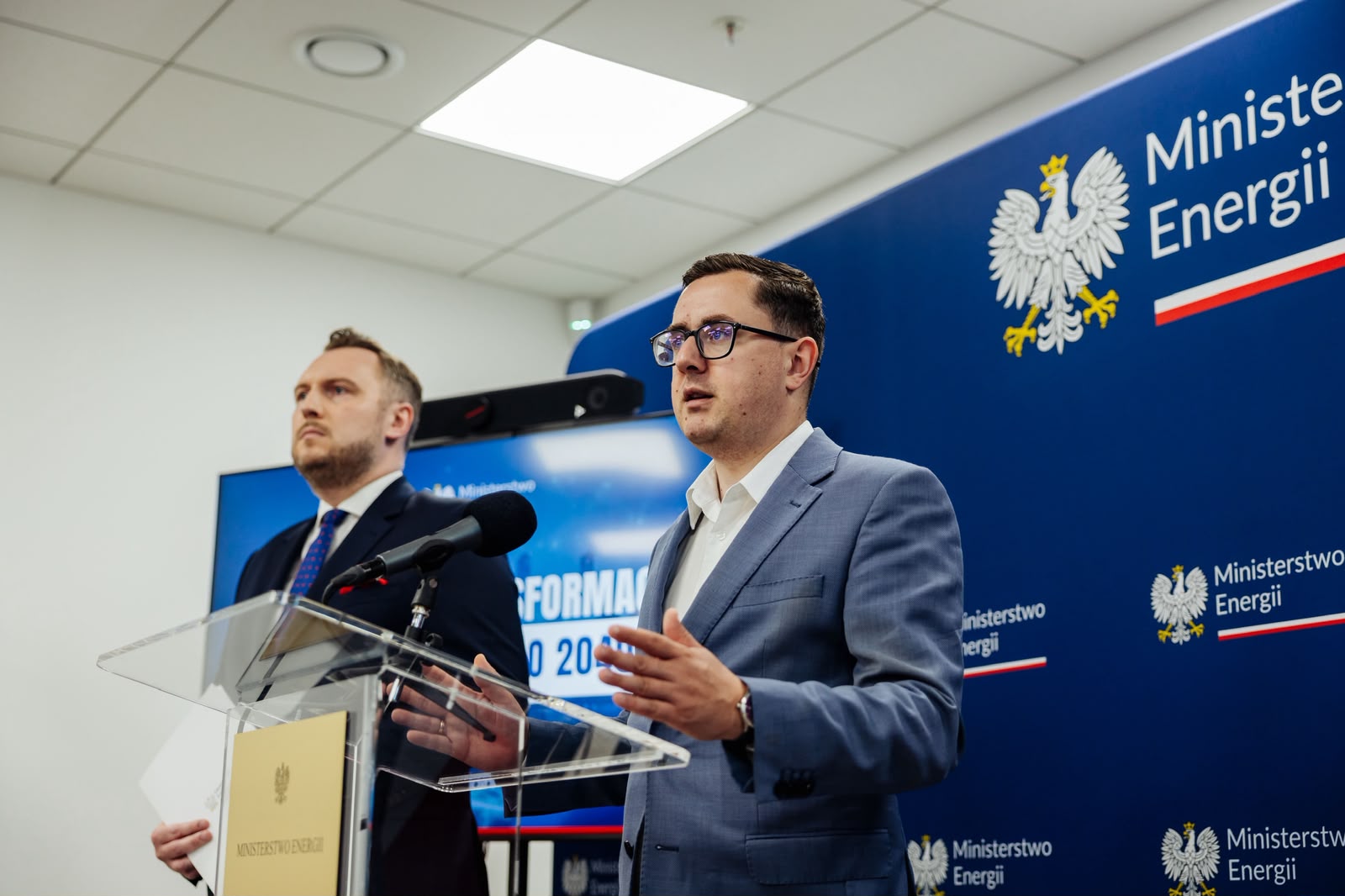 Dwaj mężczyźni przemawiający przy przezroczystej mównicy z logotypem Ministerstwa Energii podczas wystąpienia. W tle ścianka z godłem Polski i ekran z prezentacją.